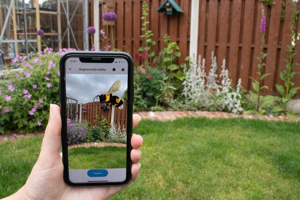 A hand holding up a mobile phone. On the screen is the 'Whats that bumblebee' app, which shows a picture of an animated bumblebee flying in a garden.