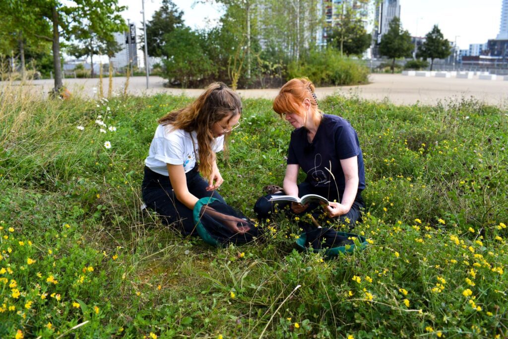 Two people sat in an area of greenspace looking at bumblebees with net and book with urban landscape