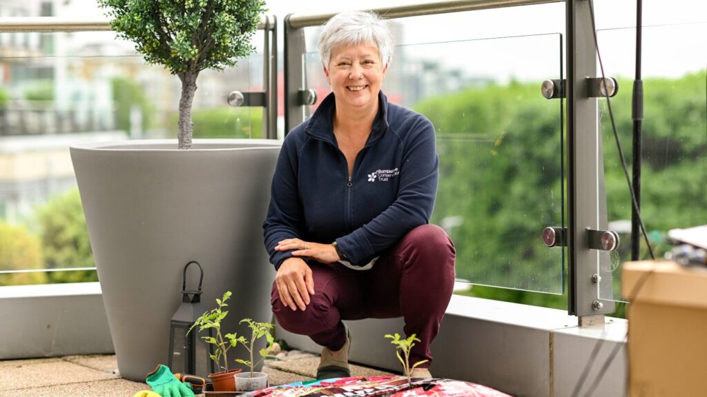 A smiling Gill crouching beside a potted plant and some gardening tools.