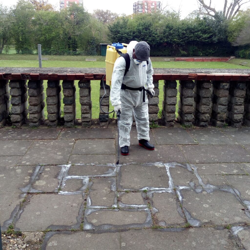 A person wearing a hazmat suit and mask spraying pesticides on a pavement.