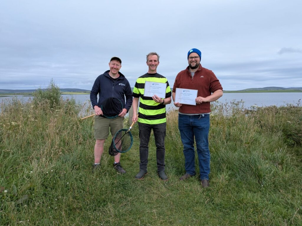 Three people holding certificates in a wildflower area